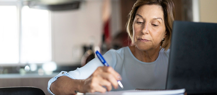 A senior woman writing while using a laptop.