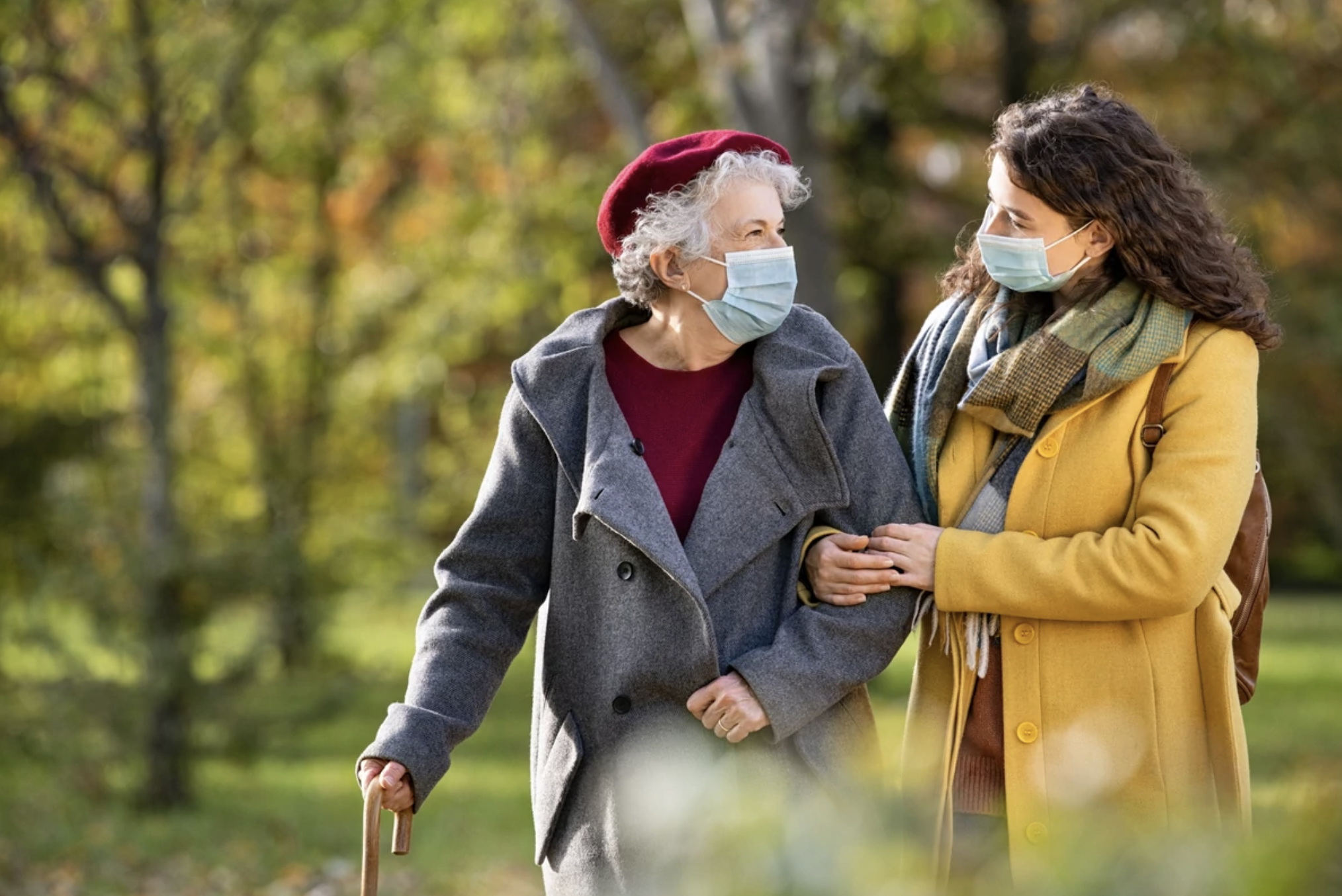 A senior woman and her caregiver taking a walk in the park during fall while wearing masks.
