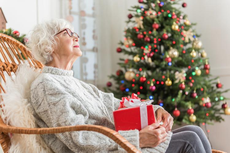 A senior woman in a chair holding a gift, with a Christmas tree in the background.