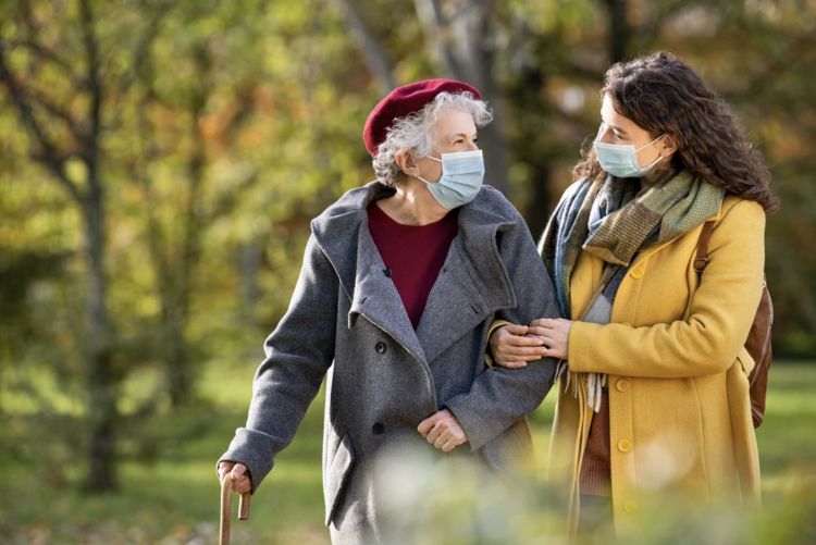 A senior woman and her caregiver taking a walk in the park during fall while wearing masks.