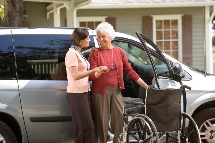 A caregiver helping a senior woman out of a wheelchair and into a car.