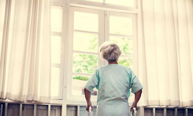 A senior woman looking out of a window while using a walking frame.